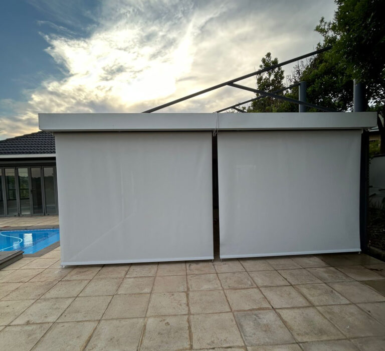 Two cream drop blinds lowered to enclose a poolside patio structure at dusk